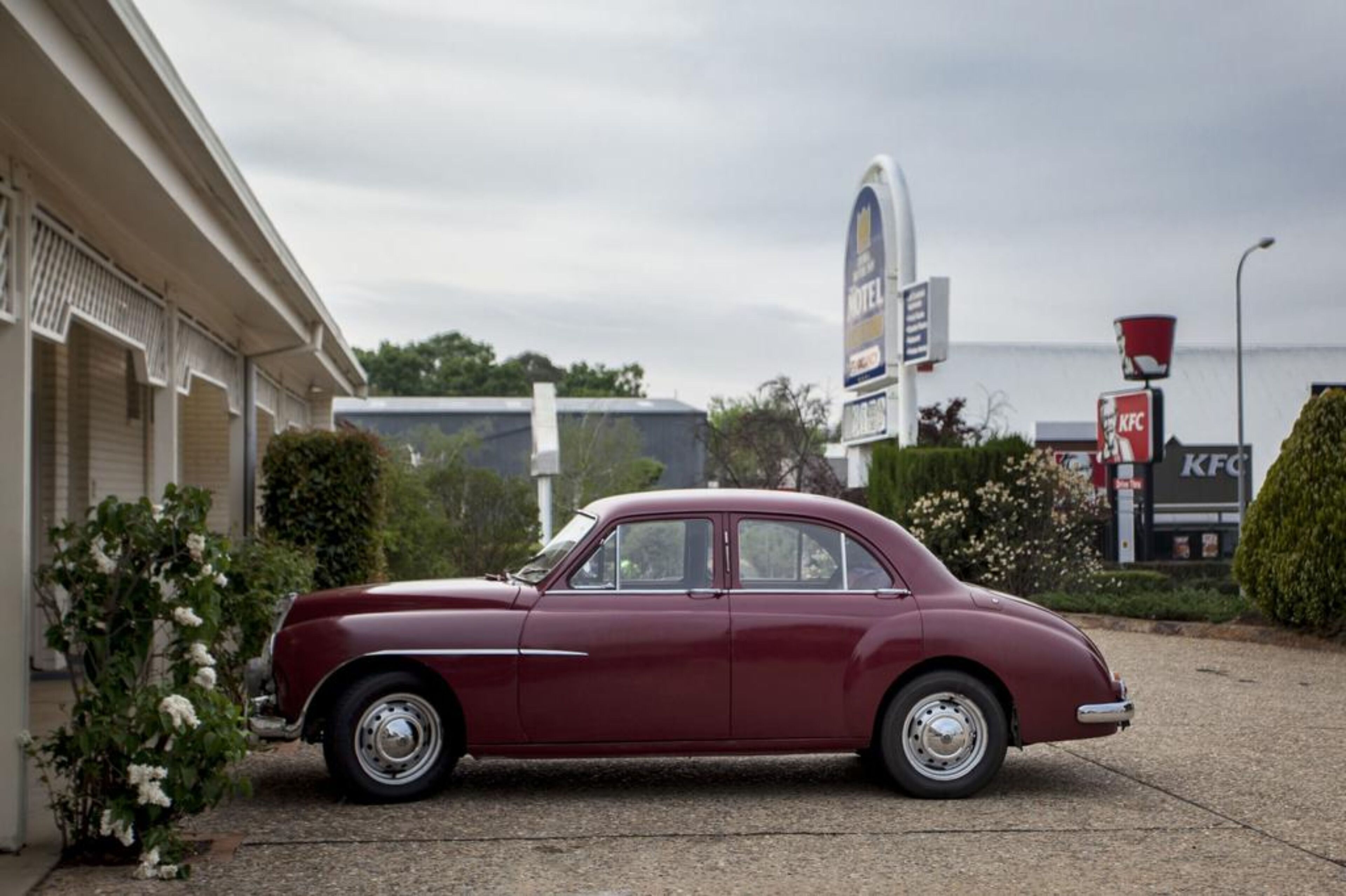 A vintage maroon car parked in front of a motel with a KFC sign in the background.