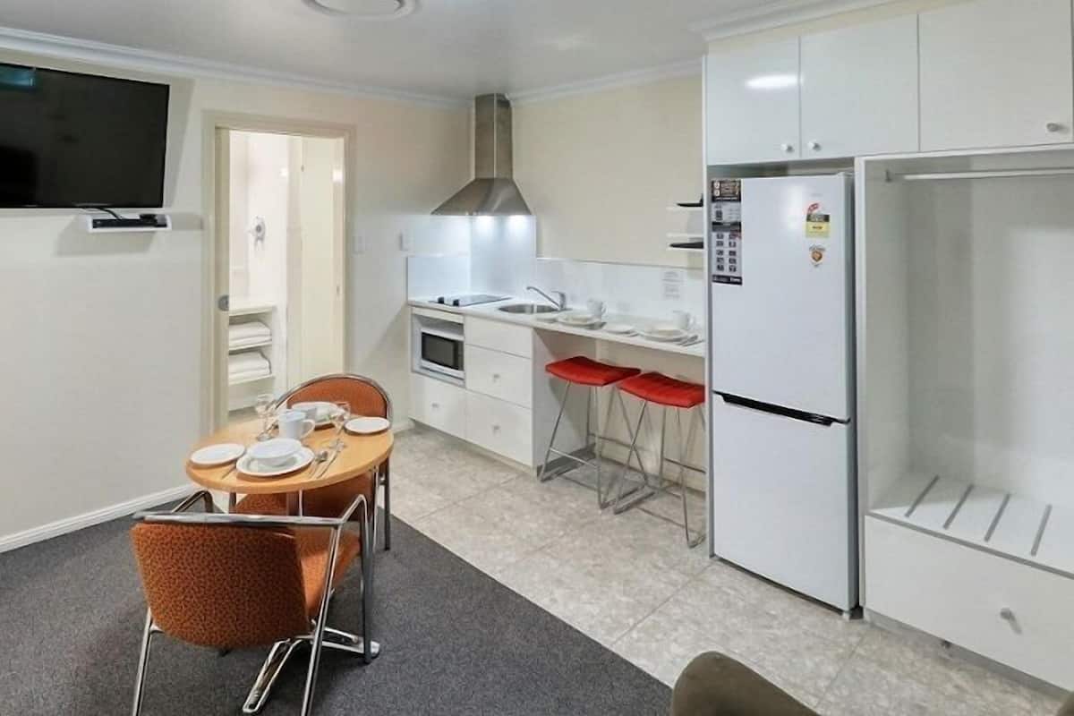 A compact kitchen with white cabinets, a built-in oven, a stainless steel range hood, and a refrigerator. There is a dining area with a round table and two chairs.