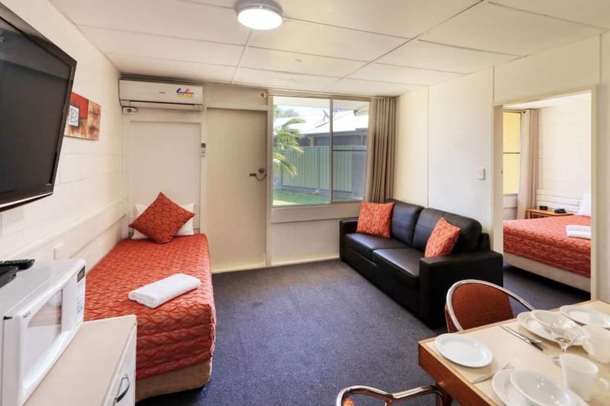 A hotel room with two single beds, a sofa, a dining table with plates and cutlery, a TV, and an air conditioning unit.