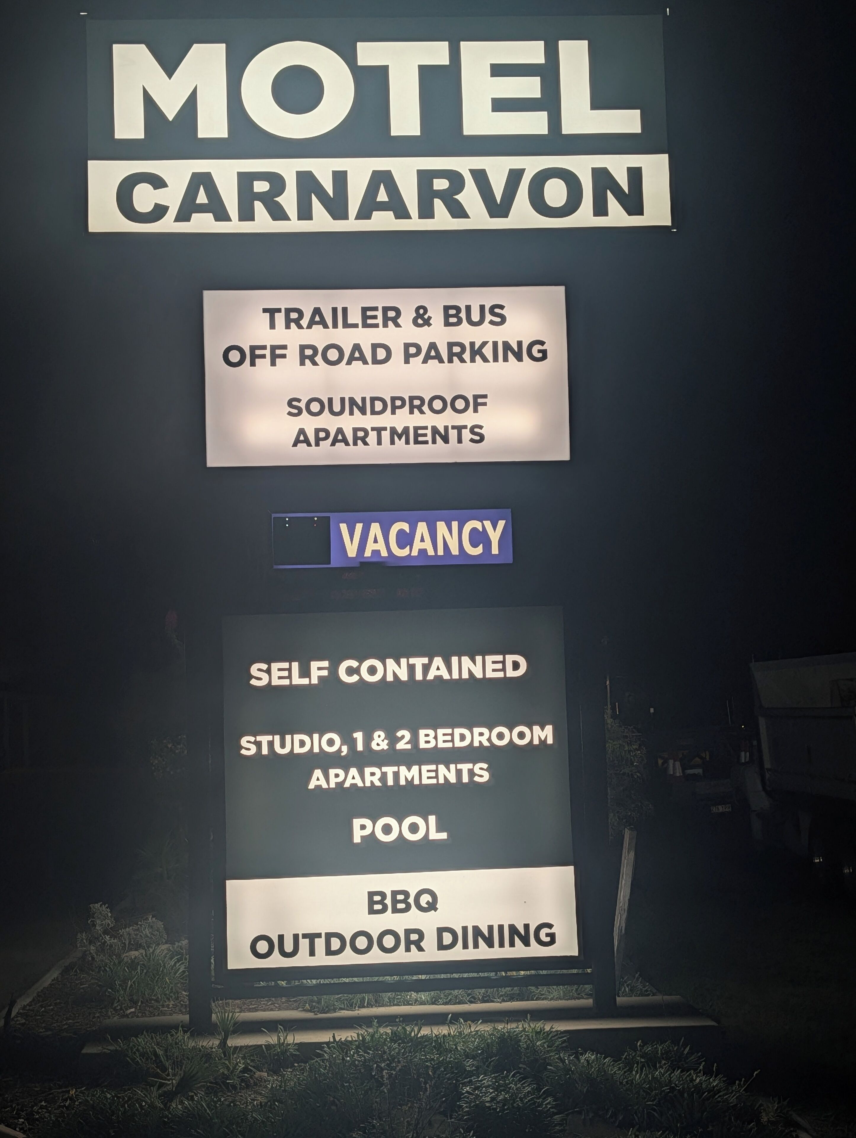 Een motelbord met 'MOTEL CARNARVON' en vermelde voorzieningen, waaronder parkeerplaats voor caravans en bussen, geluiddichte appartementen, studio's met eigen voorzieningen en 1- en 2-slaapkamerappartementen, zwembad en buitendineren bij de barbecue.