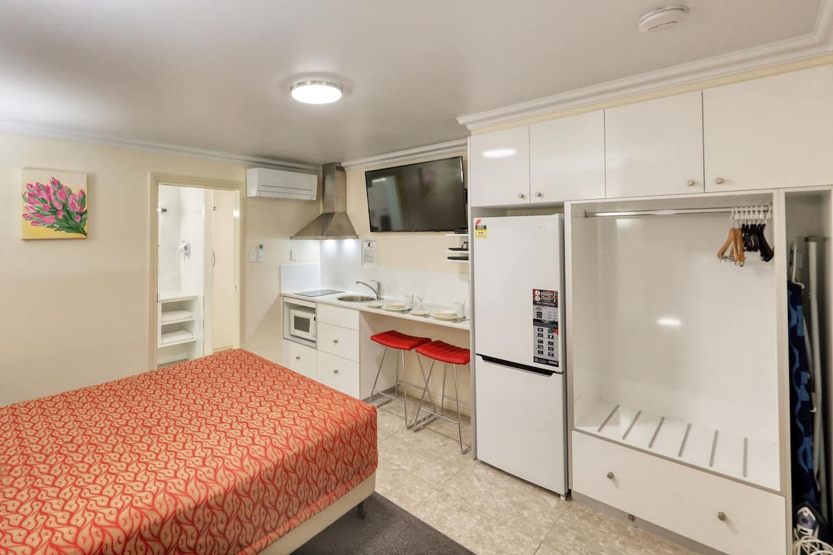 A compact hotel room with a bed, a kitchenette, a small bathroom, and a TV mounted on the wall.