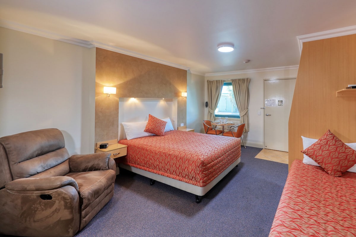 A hotel room with a bed, a sofa, a chair, a small table with a lamp, and a window with curtains.