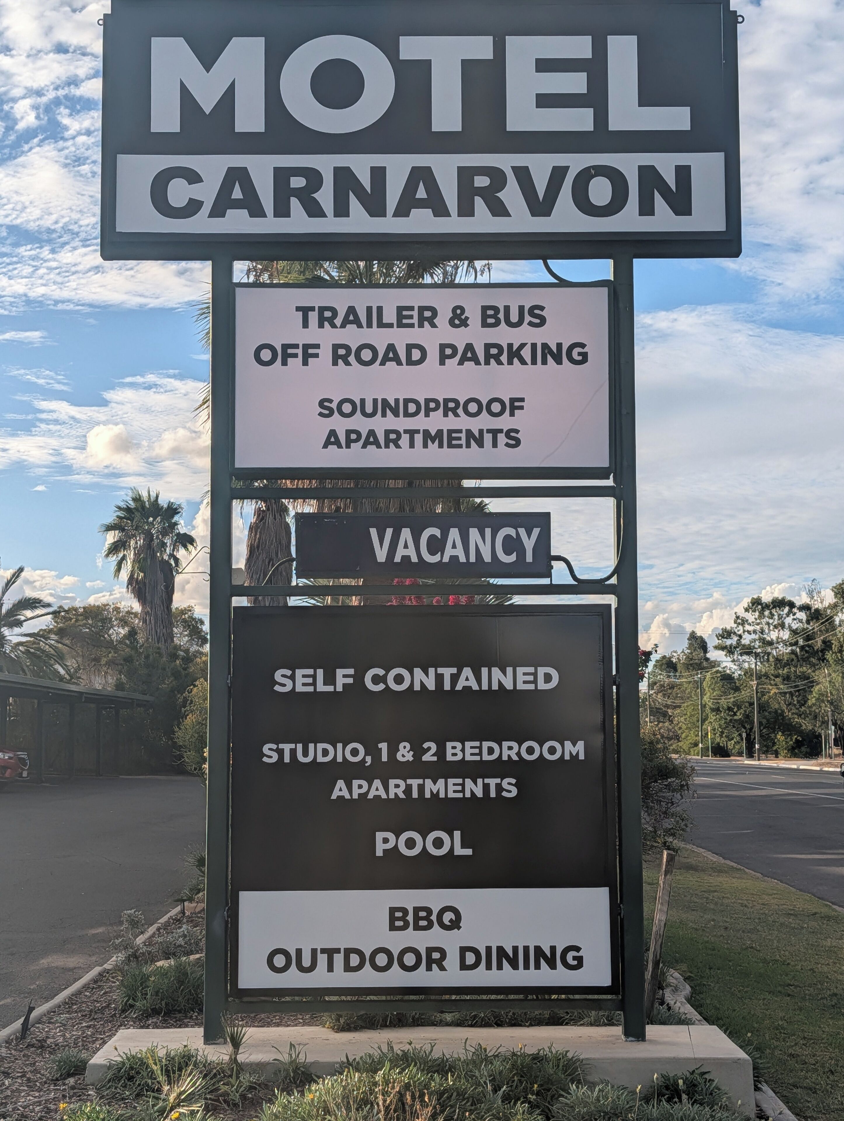 A motel sign with 'Motel Carnarvon' and amenities including trailer & bus off-road parking, soundproof apartments, self-contained studio and 2-bedroom apartments, pool, and BBQ outdoor dining.