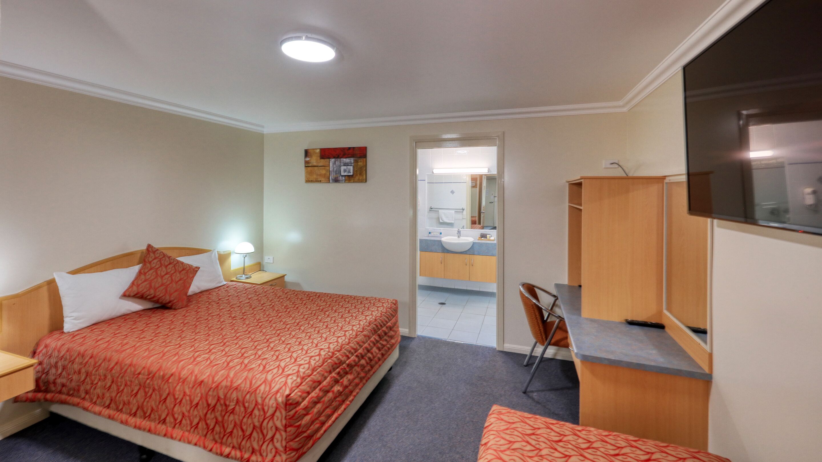 Una camera d'albergo con un letto, una scrivania, una sedia, una TV e un bagno visibile attraverso una porta aperta.
