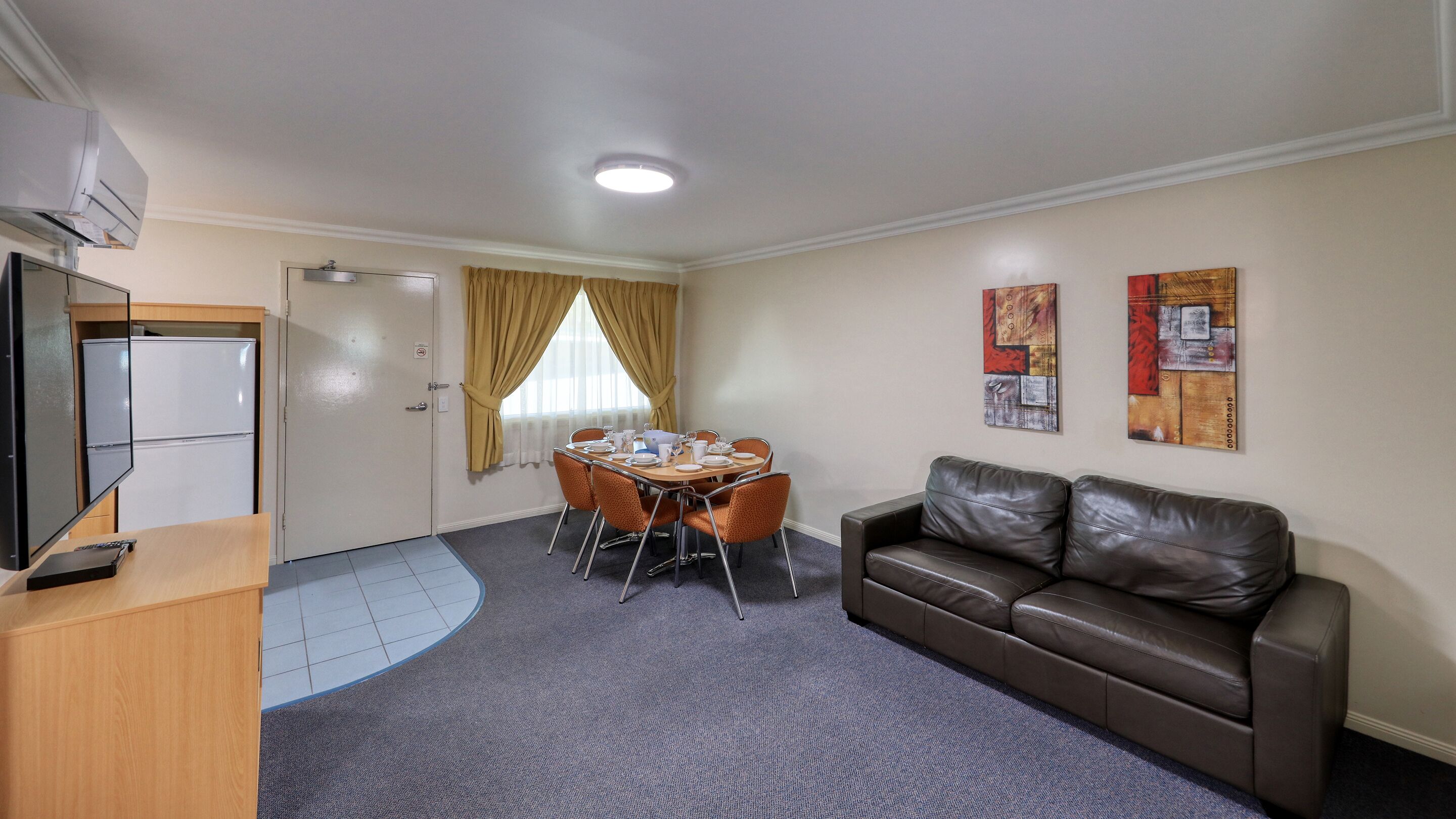 A hotel room with a dining table set for four, a leather sofa, a TV, and a refrigerator.