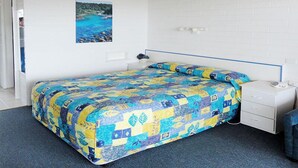 A bedroom with a bed, a nightstand, a drawer unit, and a picture of a coastal scene. - Kingfisher Motel (Merimbula)