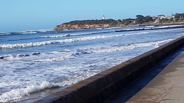 Blick auf die Küste mit einer Betonmauer, Wellen, einem Leuchtturm und Gebäuden auf einer Klippe.