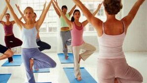 Eine Gruppe von Menschen in einem Yogakurs, stehend auf blauen Matten in einem hellen Raum mit großen Fenstern.