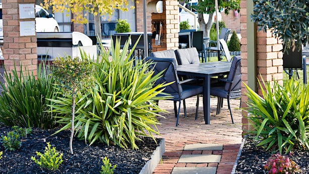 An outdoor patio area with a brick column, a table with chairs, and various plants. - Statesman Motor Inn (Corowa)
