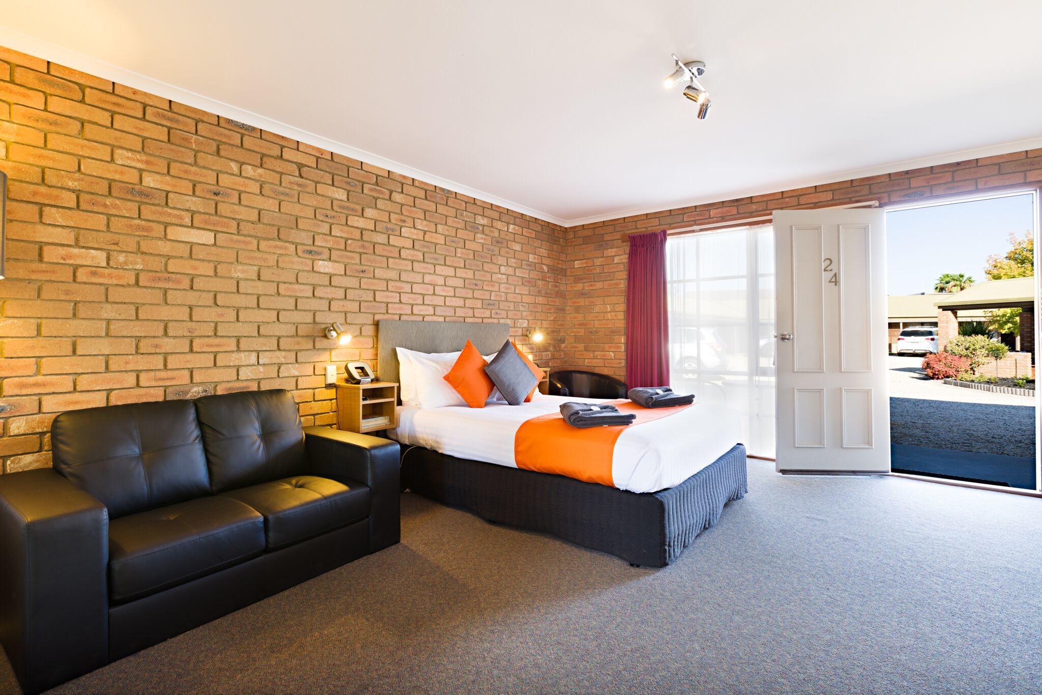 Ein Hotelzimmer mit Backsteinwand, einem Bett mit orangefarbener Bettwäsche, einer schwarzen Ledersofa und einer Schiebetür zum Außenbereich.