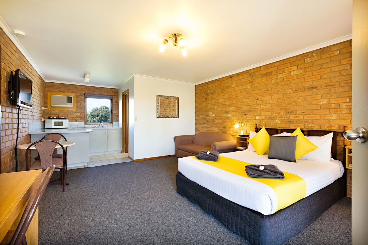 A hotel room with a bed, a desk, a chair, a kitchenette, and a brick wall.