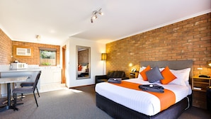 A modern hotel room with a brick wall, a large bed with orange bedding, a desk with a chair, and a small kitchen area. - Statesman Motor Inn (Corowa)