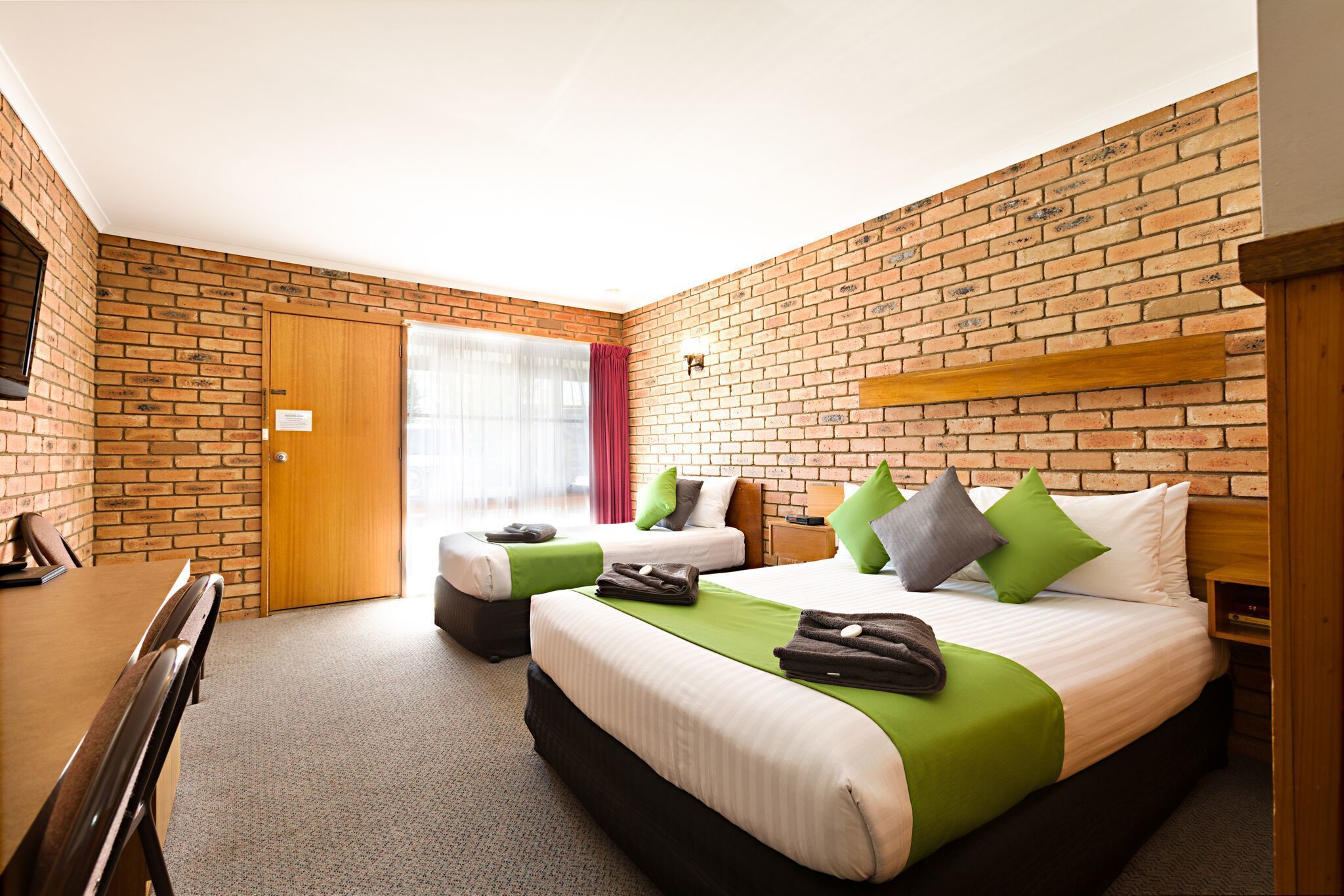A hotel room with two beds, a wooden desk, and a brick wall.
