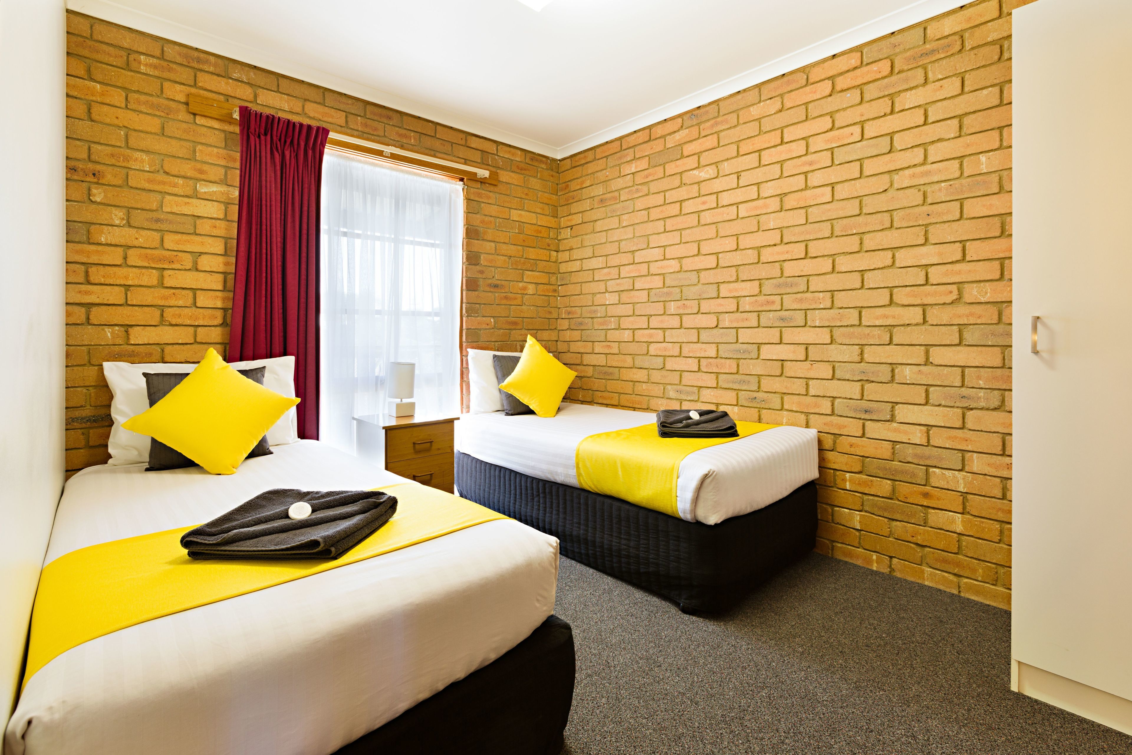 Ein Hotelzimmer mit zwei Betten, einer Ziegelwand, einem mit Vorhängen versehenen Fenster und einem Teppichboden.