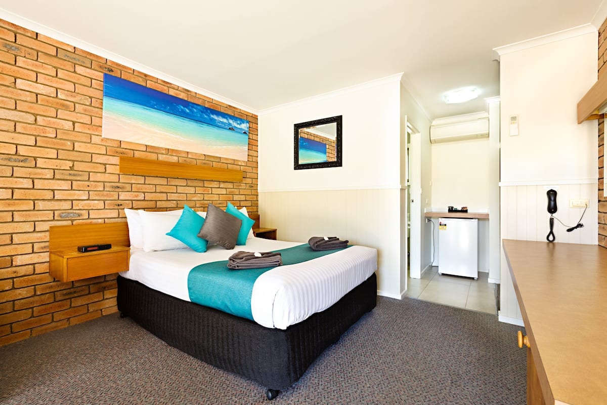A hotel room with a brick wall, a bed with white bedding and teal pillows, a small bedside table with a book, a painting of a beach, a mirror, a kitchenette, and a telephone.
