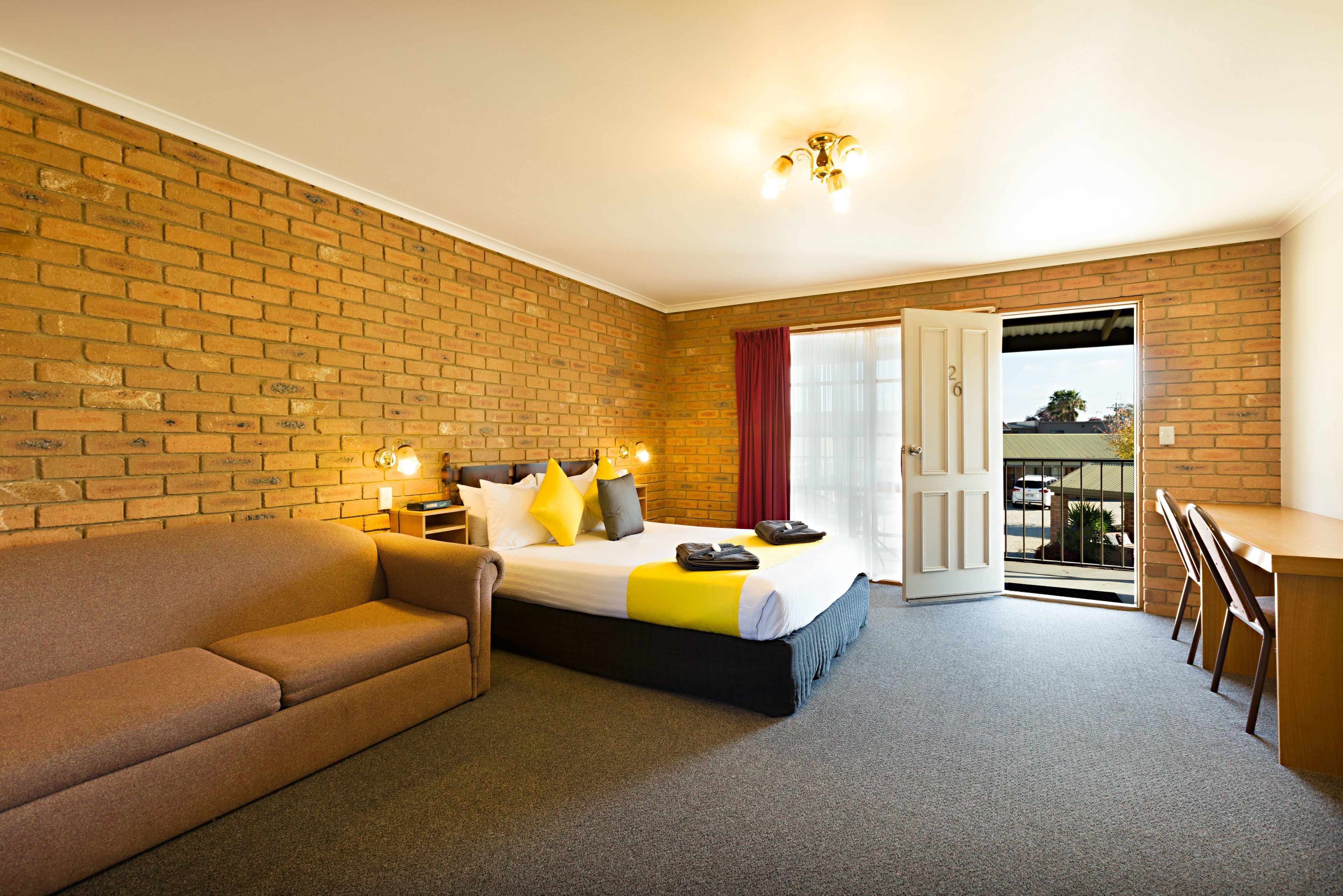 A hotel room with a brick wall, a bed with a yellow and white bedspread, a brown sofa, a desk, and a balcony door.