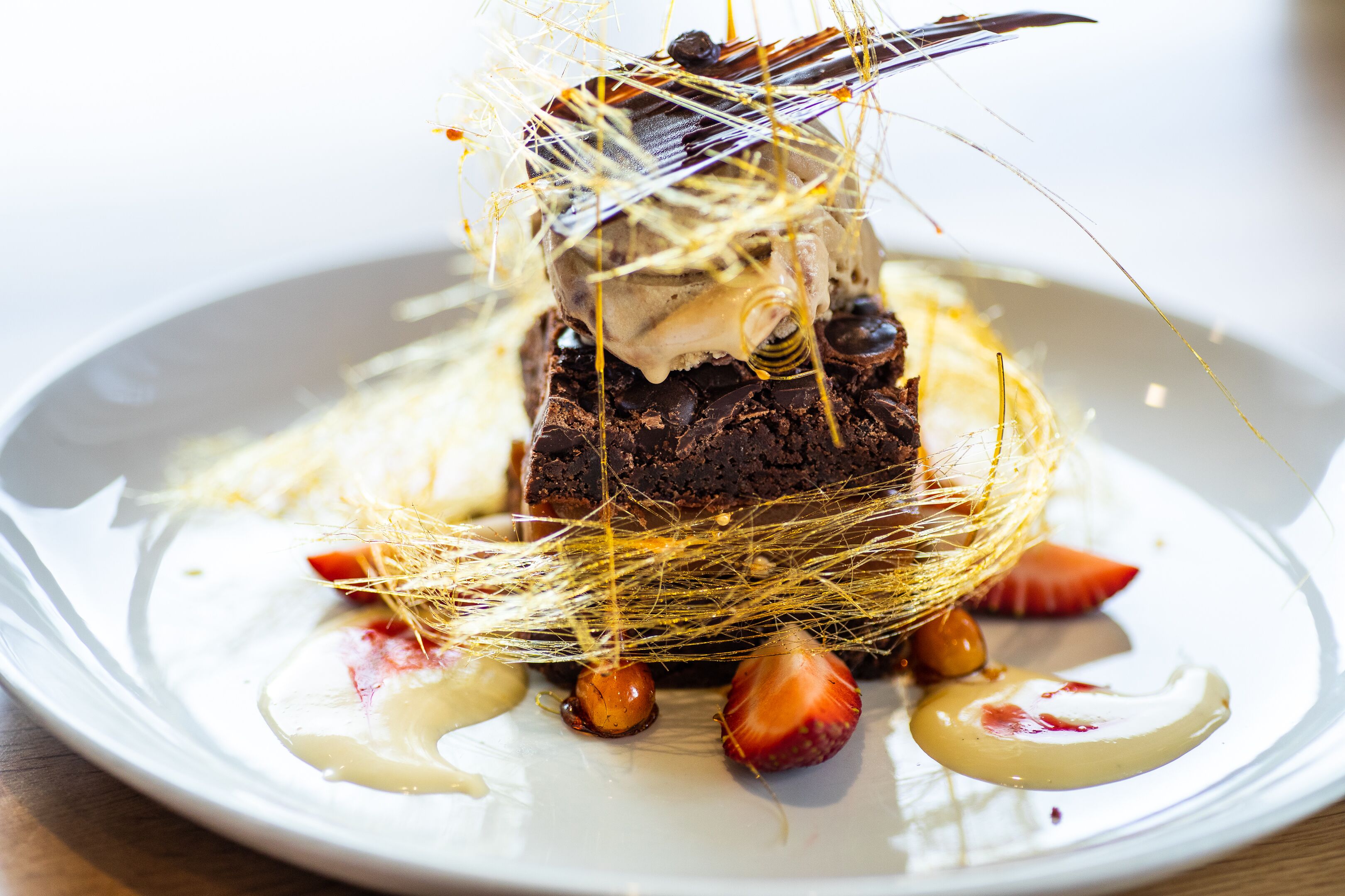Un dessert con cioccolato, fragole e un nido decorativo di fili dorati su un piatto bianco.