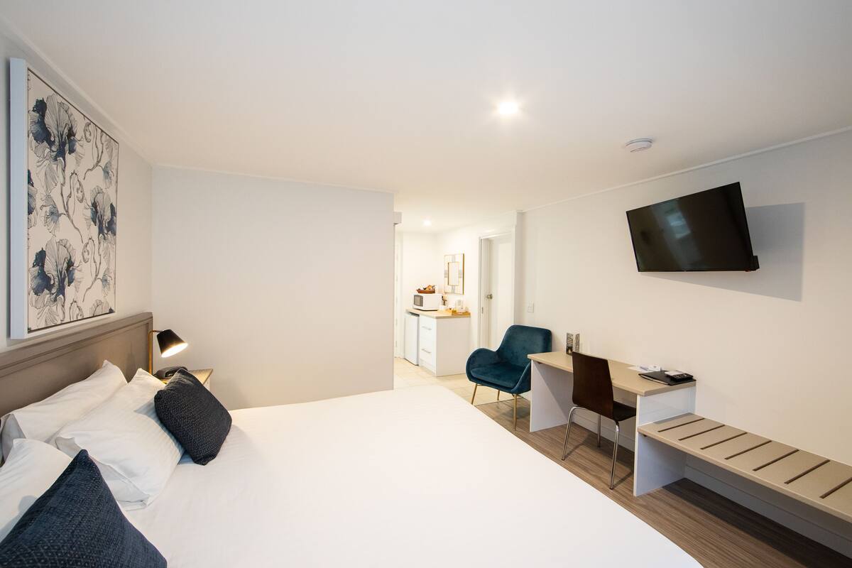 A modern hotel room with a large bed, a desk, a chair, a TV, and a small kitchenette in the background.