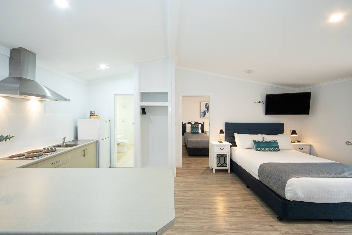A modern hotel room with a kitchenette, a bed with a headboard, bedside tables, and a flat-screen TV mounted on the wall.