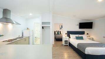 A modern hotel room with a kitchenette, a bed with a headboard, bedside tables, and a flat-screen TV mounted on the wall.
