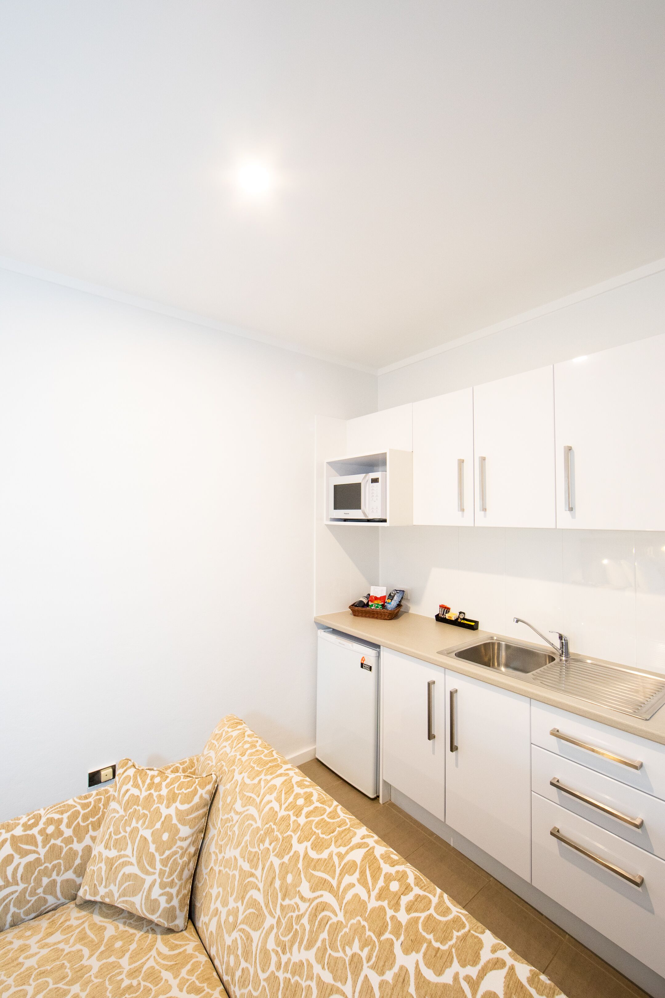 A compact kitchen with white cabinets, a microwave, a sink, and a small refrigerator. There is a beige patterned sofa in the foreground.