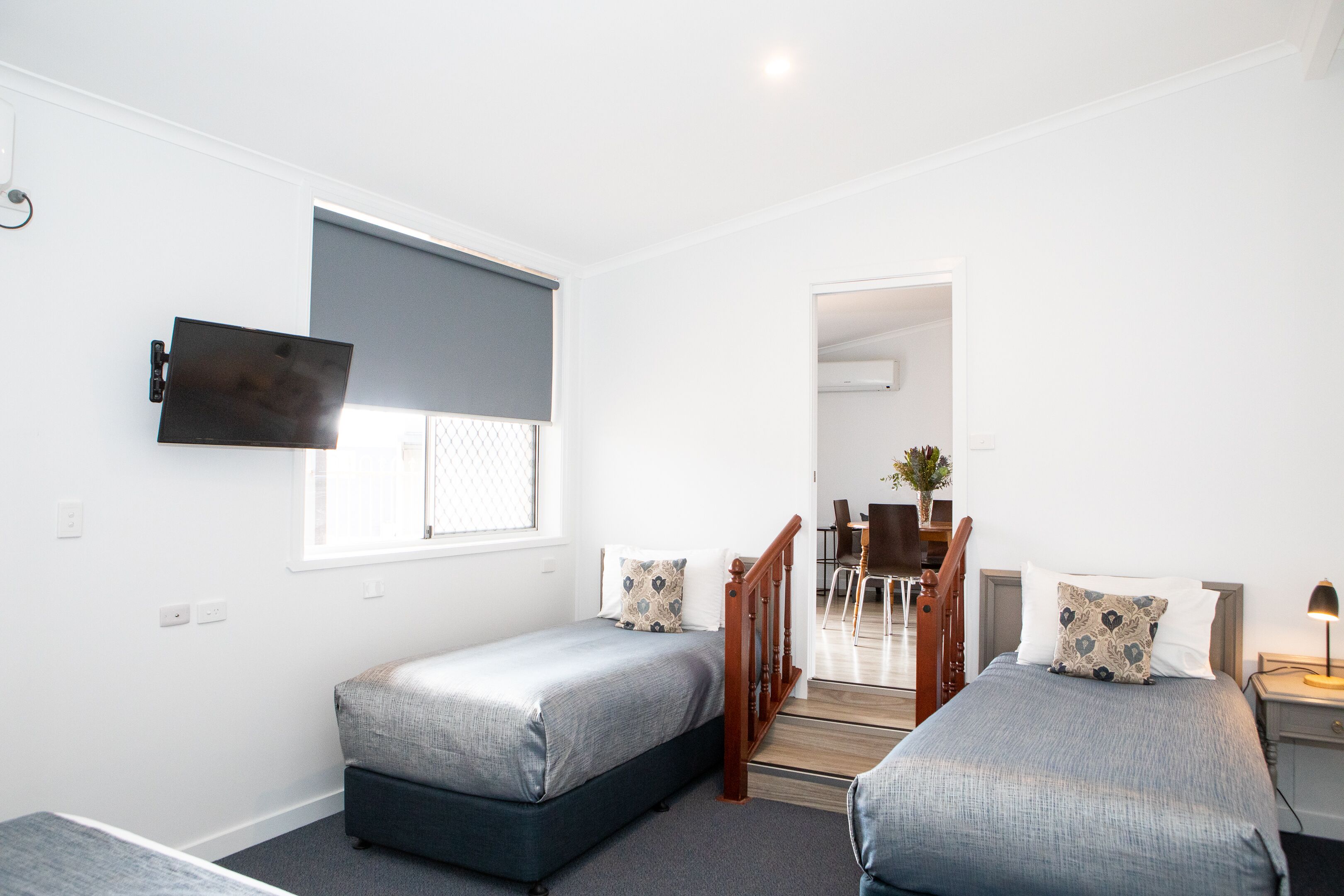 A bedroom with two single beds, a TV mounted on the wall, a desk with a chair, and a staircase leading to another room.