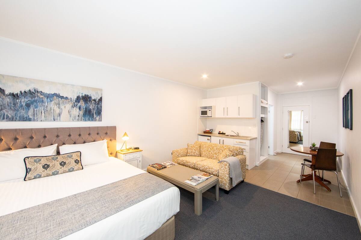 A modern hotel room with a bed, a sofa, a coffee table, a desk, and a kitchenette.