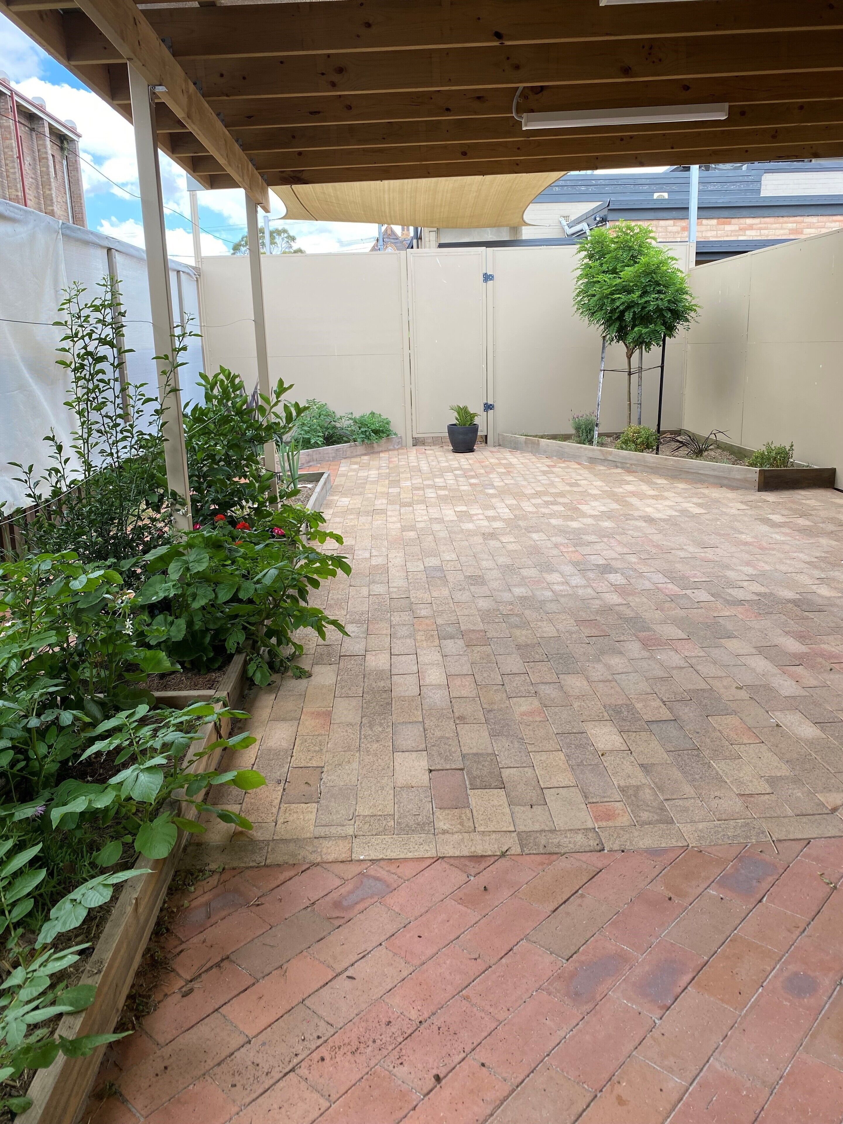 A covered patio area with a brick floor, surrounded by plants and a wooden fence.