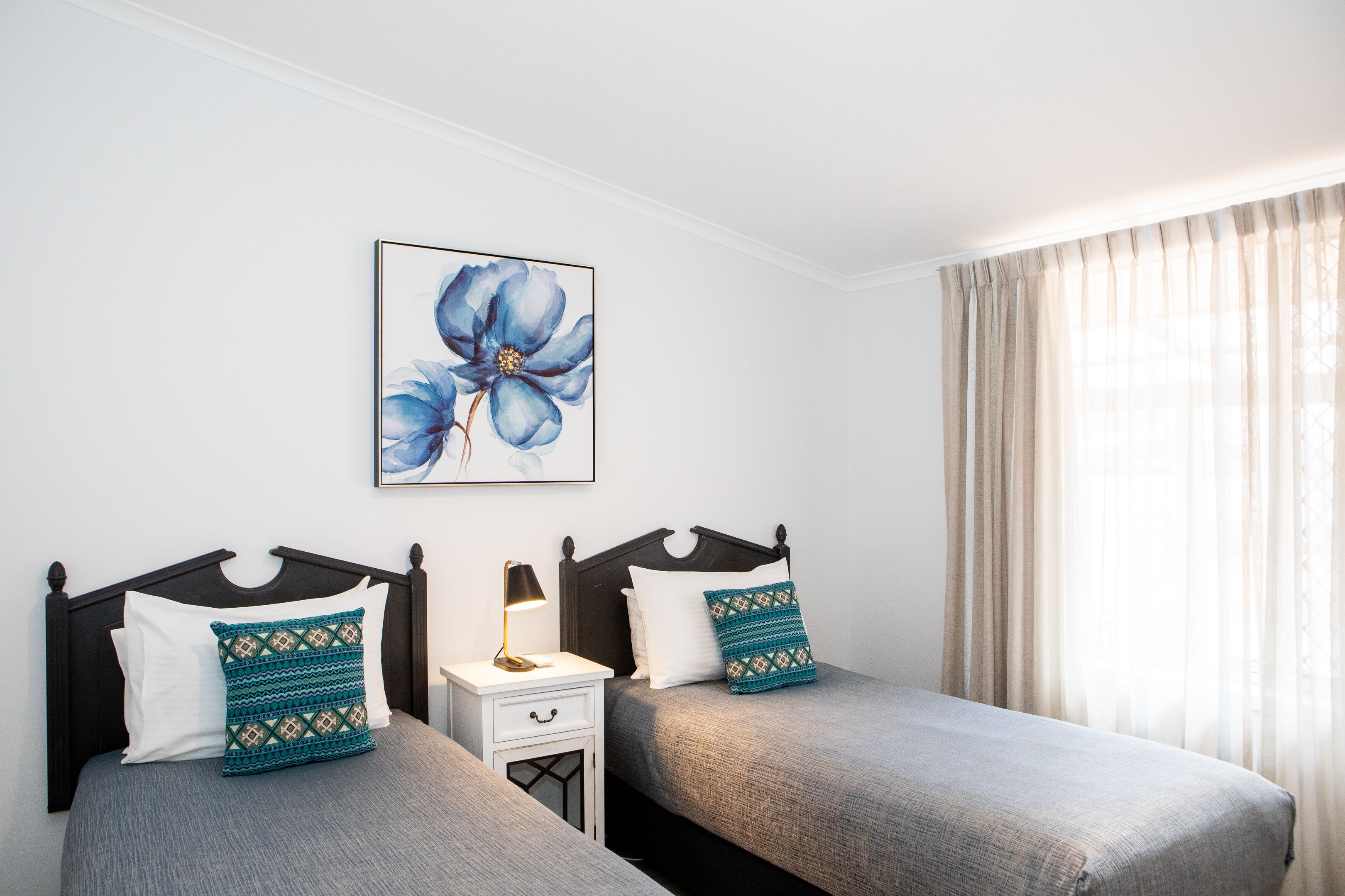 Un dormitorio con dos camas, una mesita de noche, una lámpara, una obra de arte floral enmarco y cortinas de gasa.