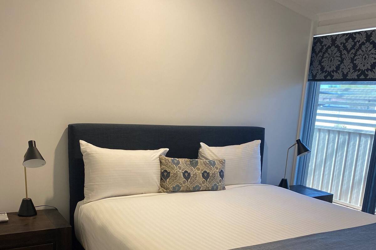 A neatly made bed with white linens and a grey throw, a dark headboard, and a bedside table with a lamp.