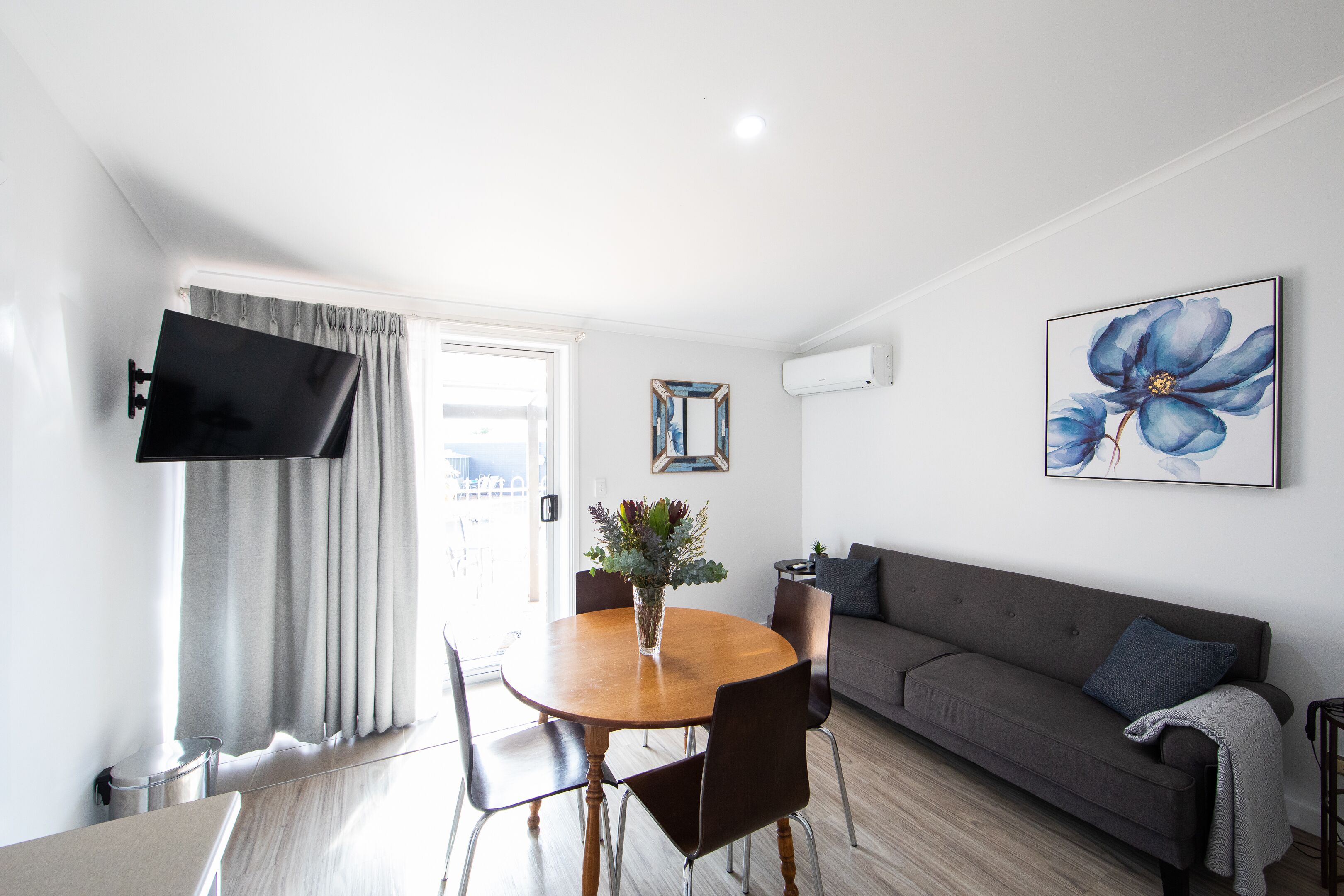 Una sala de estar moderna con una mesa de comedor redonda de madera, un sofá gris y un televisor de pantalla plana montado en la pared.