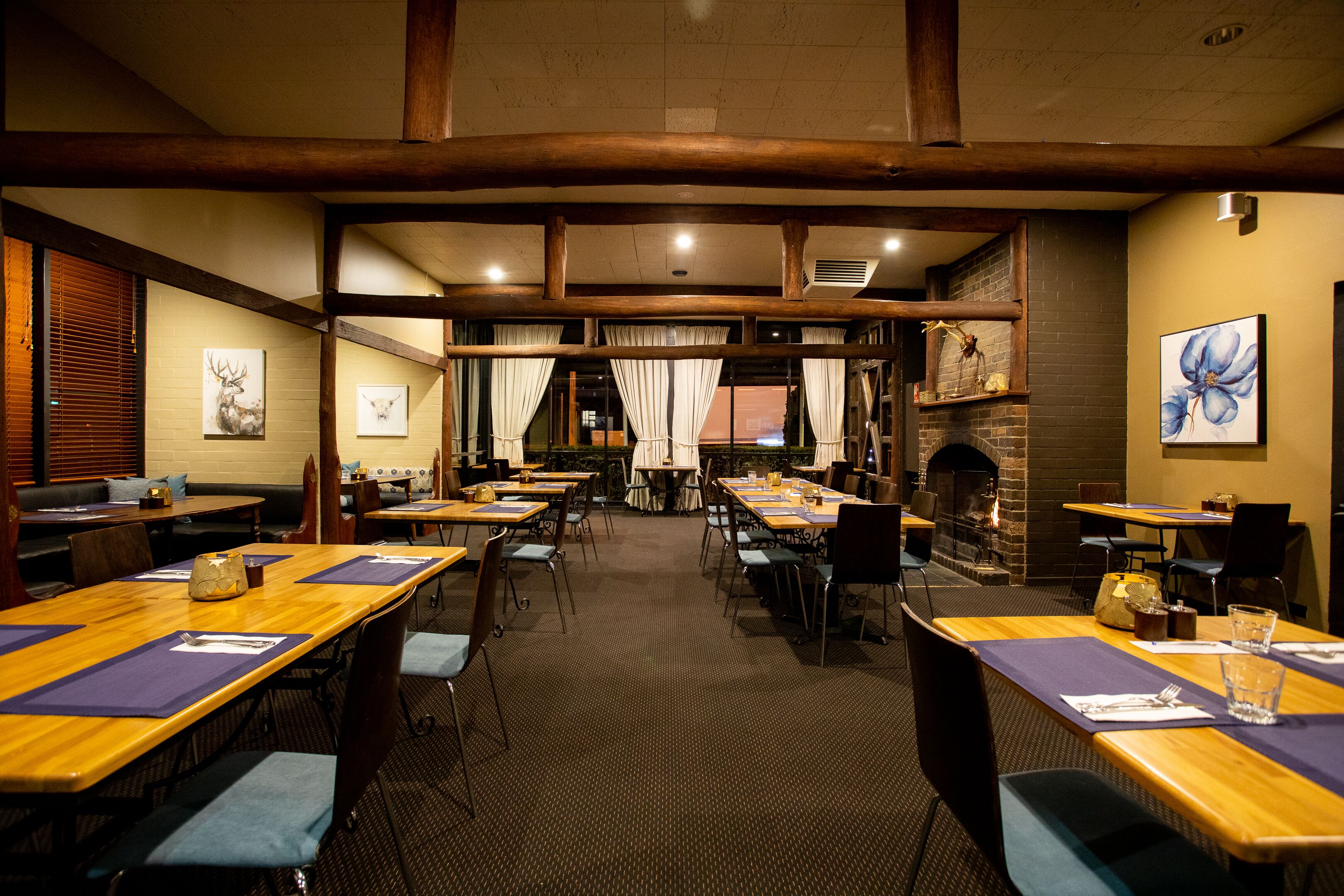 A restaurant with multiple tables and chairs, wooden beams on the ceiling, and a brick wall with a fireplace.