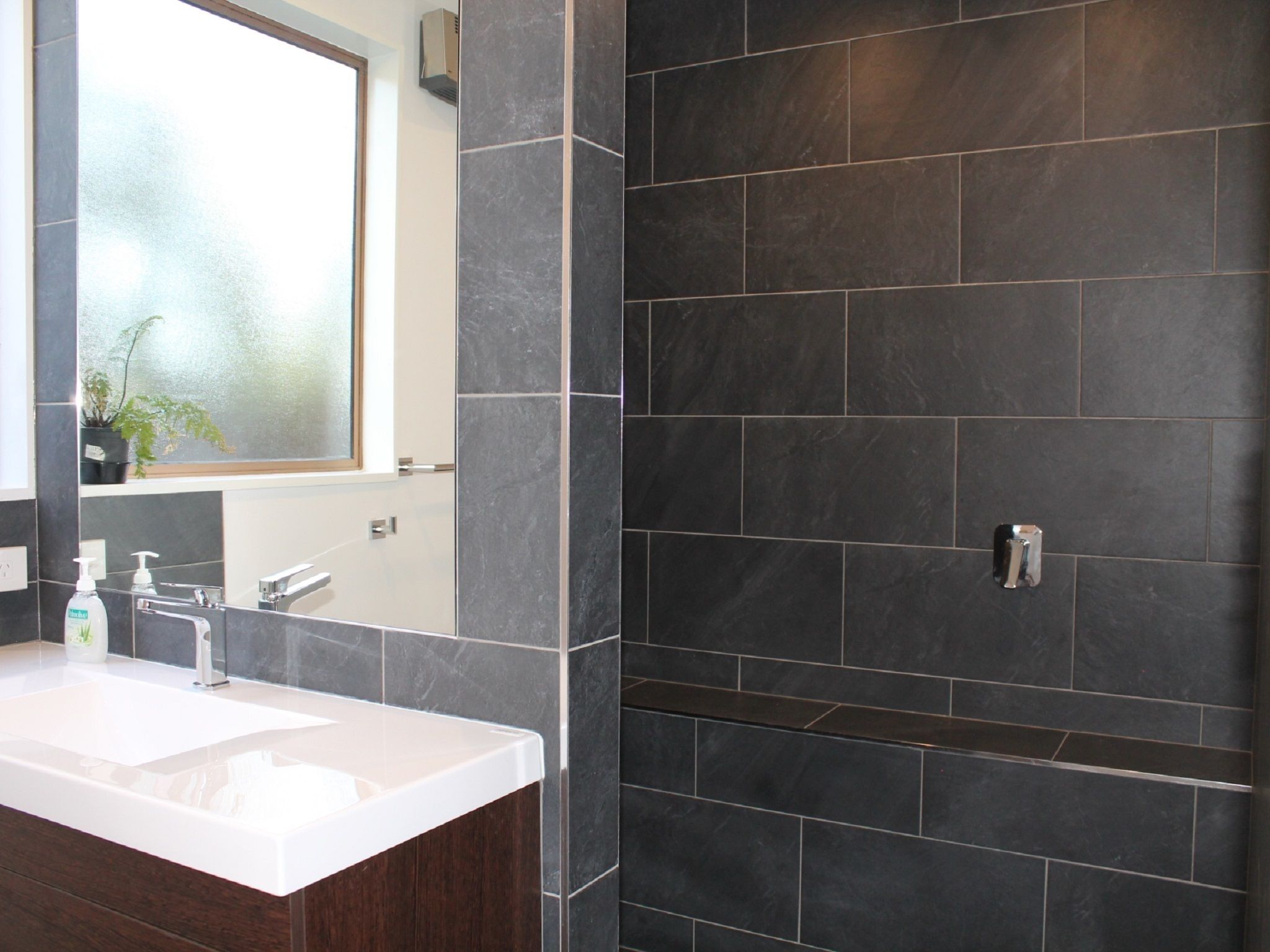 Un baño moderno con un lavabo blanco, baldosas oscuras y una ventana con vistas.