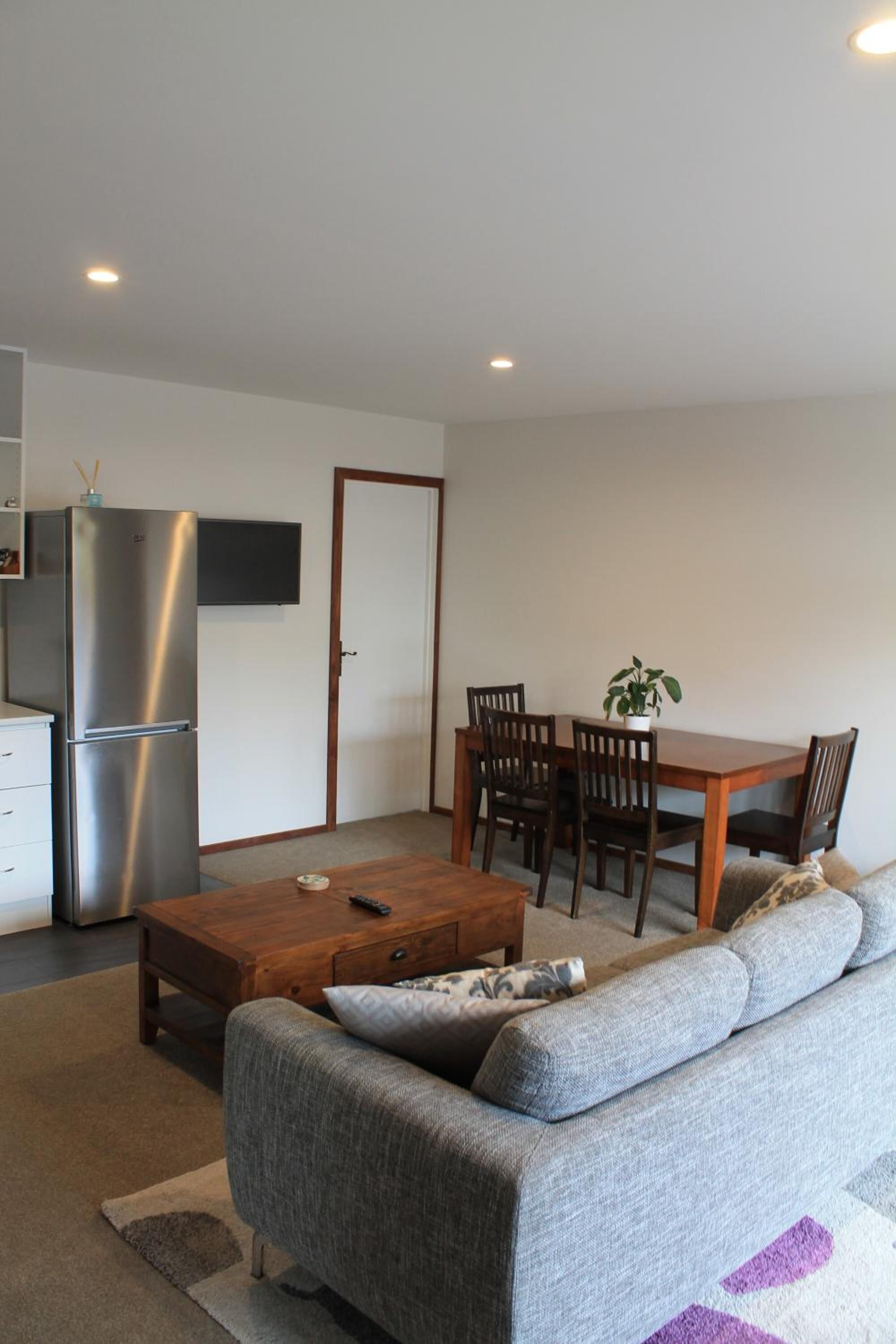 Ein modernes Wohnzimmer mit grauem Sofa, Holzesstisch und Stühlen. Ein Couchtisch steht in der Mitte. Ein Edelstahlkühlschrank steht an der Wand.