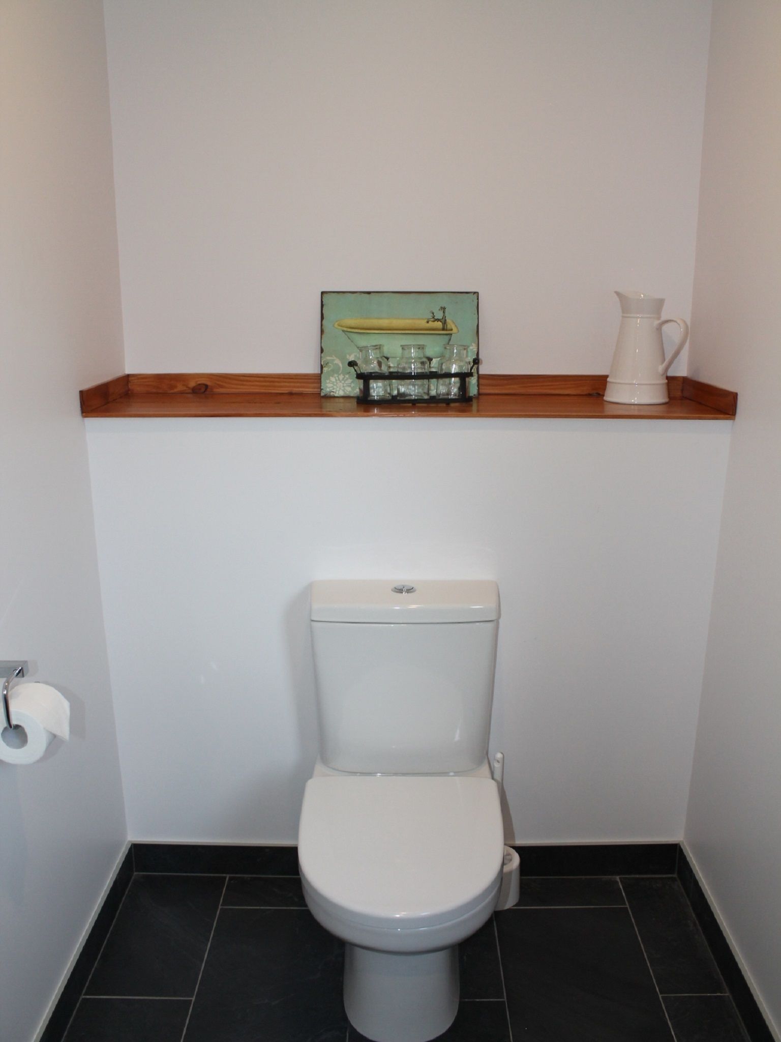 Een badkamer met een toilet, een plankje met decoratie en een witte kan.