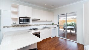 A modern kitchen with white cabinets, a microwave, a gas stove, and a dishwasher. There is a sliding glass door leading to an outdoor patio area with a table and chairs. - Canberra Ave Villas (Queanbeyan)