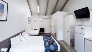 A hotel room with a bed, a desk, a chair, a television, and an air conditioning unit. - Canberra Ave Villas (Queanbeyan)
