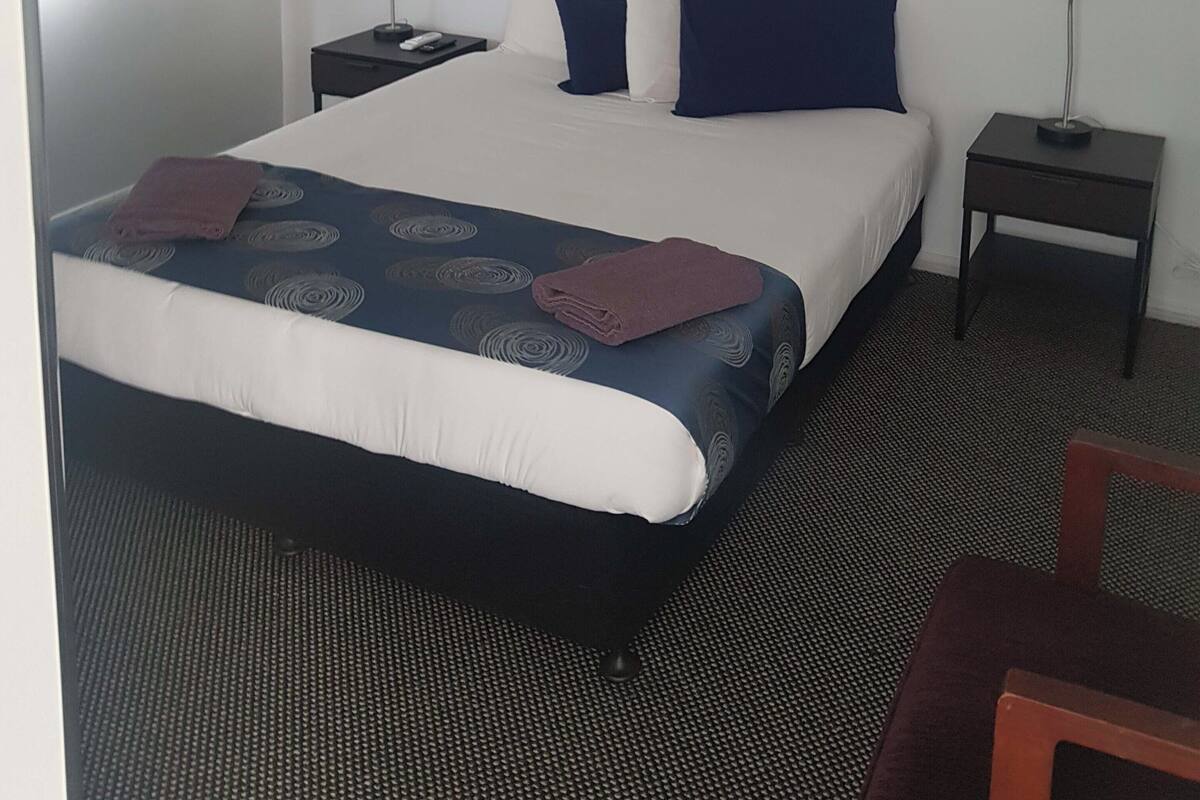 A hotel room with a bed, two bedside tables, a lamp, a framed picture of a parrot, and a chair.