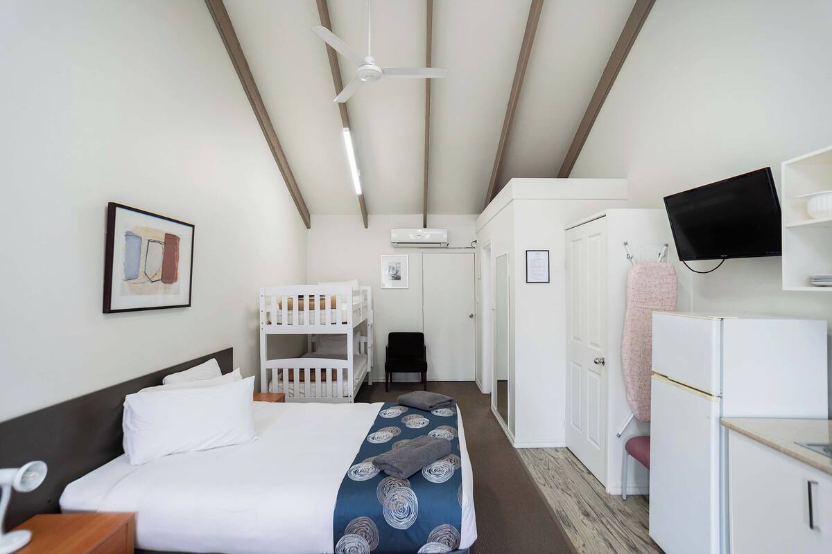 A hotel room with a bed, a ceiling fan, a TV, a desk, a chair, a wardrobe, and a kitchenette.