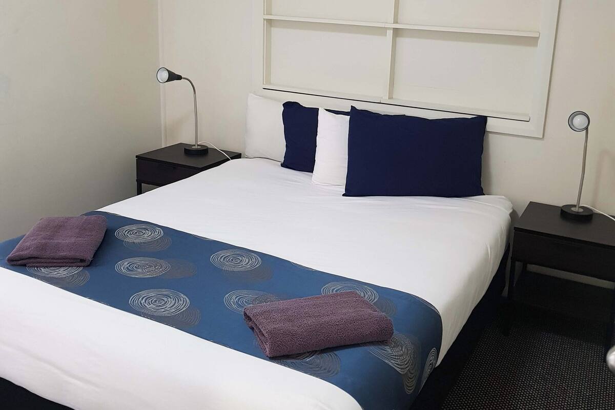 A hotel room with a large bed, two bedside tables, a wall-mounted shelf, and a small desk.