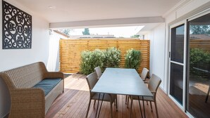 An outdoor patio with a wooden deck, a wicker sofa, a dining table with chairs, and a wooden fence in the background. - Canberra Ave Villas (Queanbeyan)