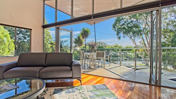 Una sala de estar moderna con un sofá de cuero, una mesa de centro de vidrio y un amplio ventanal corredero que da a un patio exterior con vistas a árboles y un cielo despejado.