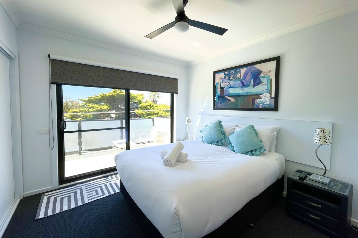 A hotel room with a bed, a ceiling fan, a painting, a nightstand, and a view of the outdoors.