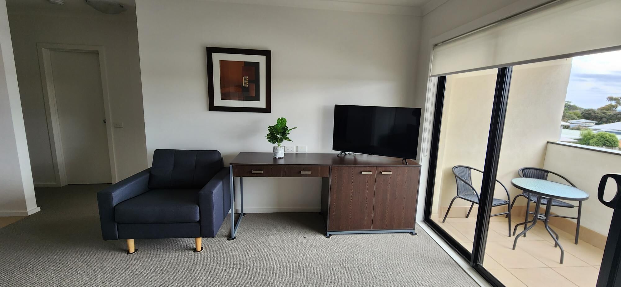 A room with a dark blue armchair, a wooden desk with a TV, a framed picture on the wall, and a balcony with chairs and a table.