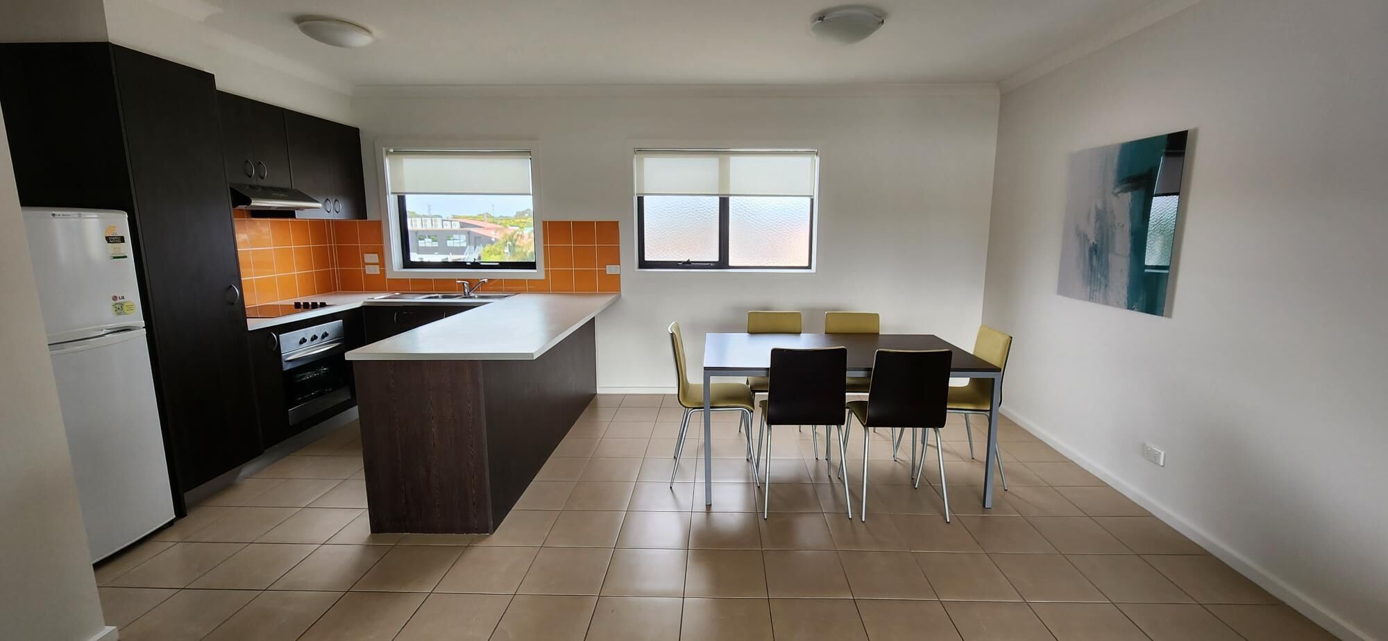 A modern kitchen with a central island, a dining area with a table and chairs, and two windows providing natural light.