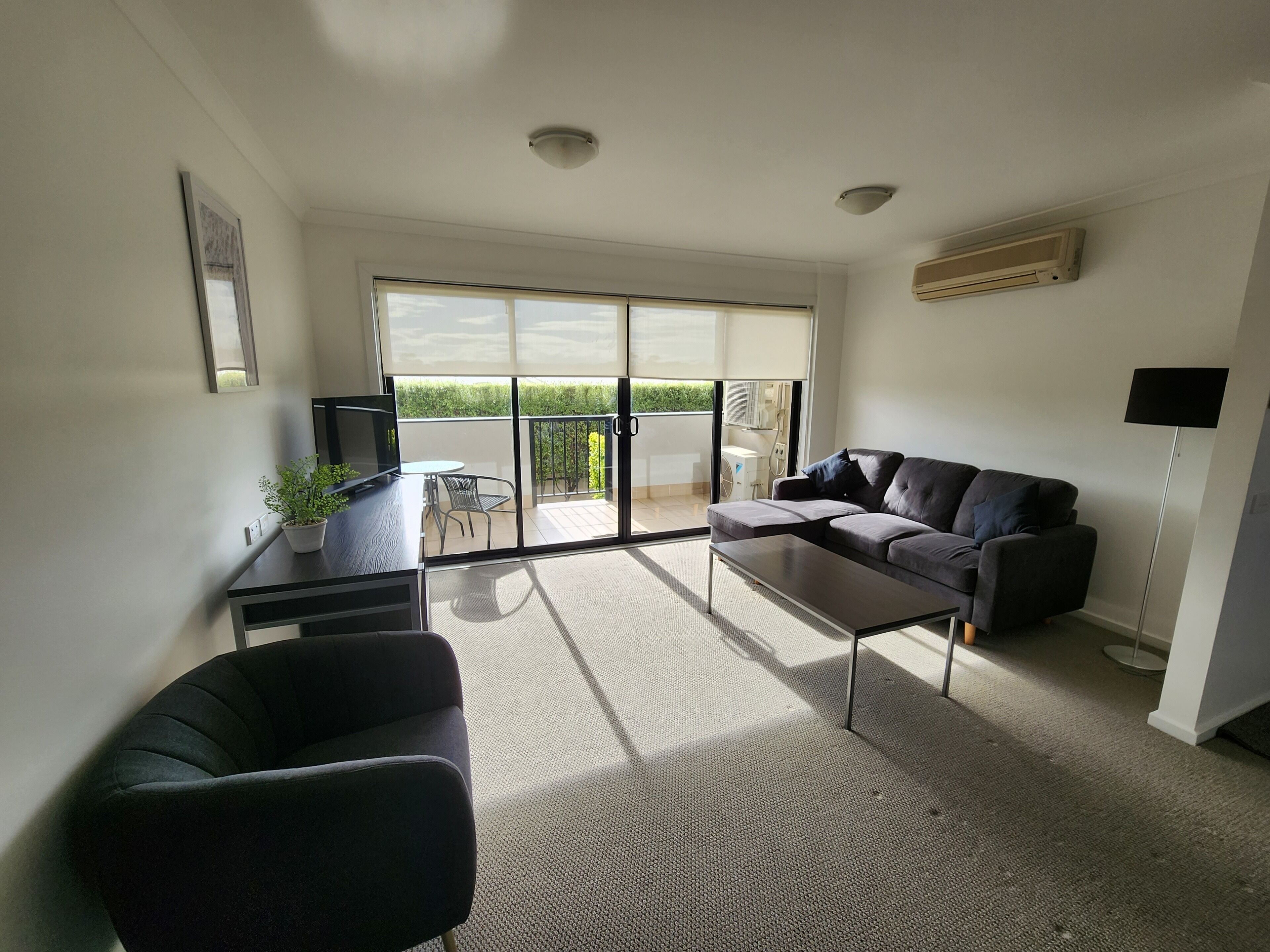 A modern living room with a sofa, a coffee table, and a TV on a stand. There is a balcony with a view of greenery.