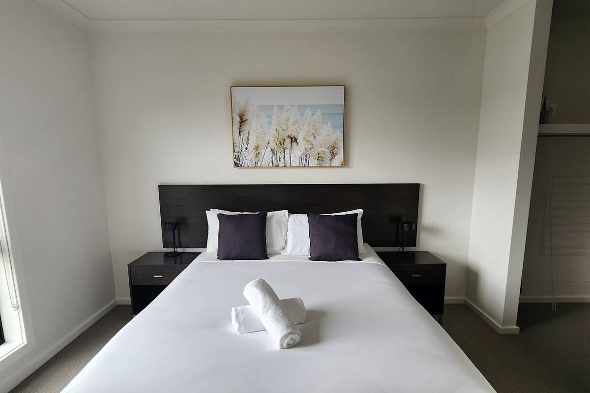 A large bed with white bedding, a dark headboard, and a painting of pampas grass on the wall.