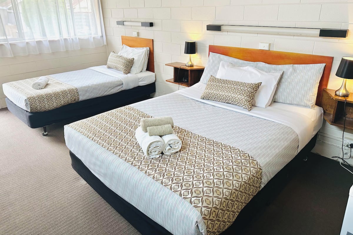 Habitación de hotel con dos camas, cada una con cabecero de madera, ropa de cama blanca y almohadas estampadas. Hay mesitas de noche con lámparas y una ventana con cortinas traslúcidas.