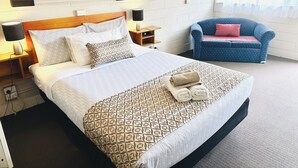 Habitació d'hotel amb un llit, una tauleta de nit, una làmpada, un otomà blau i una finestra amb cortines.