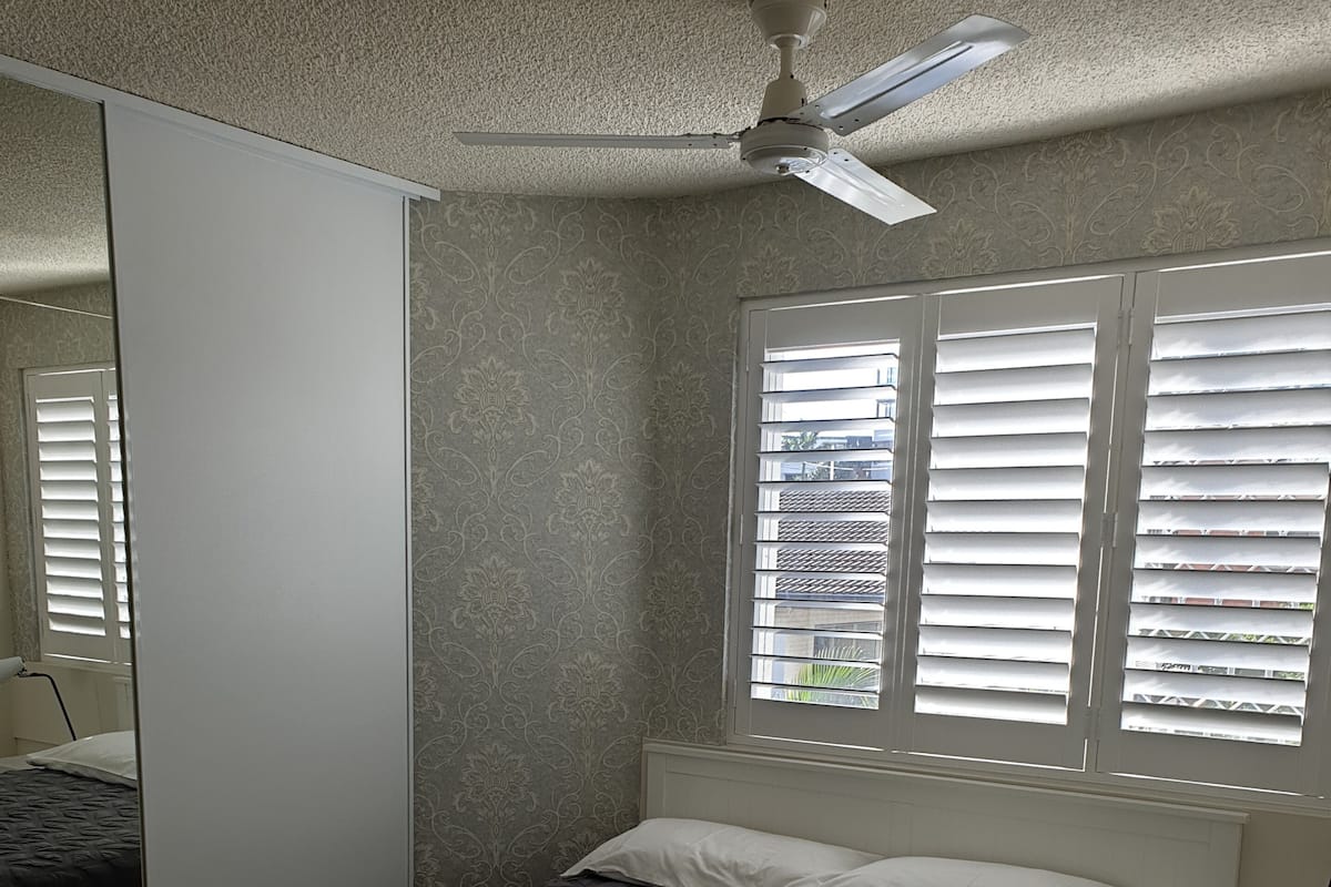 Un lit bien fait, recouvert d’une couverture à motifs sombres, avec une serviette pliée et un petit flacon de lotion posé dessus. Au-dessus du lit, on aperçoit un ventilateur de plafond, et la pièce est dotée d’une fenêtre avec des stores et d’une table de chevet sur laquelle se trouve une lampe.